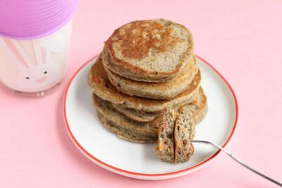Image of a stack of small, perfectly browned, wheat-colored, carrot pancakes sitting on a small white ceramic plate with an orange trim. The plate rests on a solid pastel pink background next to glass with an image of a two dimensional bunny face on it.